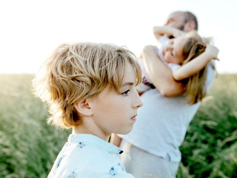 Trauriger Junge steht im Vordergrund des Bildes, im Hintergrund spielt der Vater mit der Schwester.