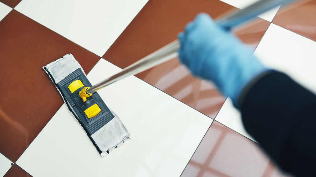 Eine Hand hält einen Mopp und wischt braun weiß karierte Fliesen.