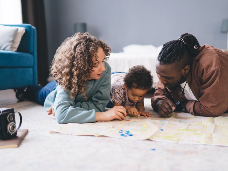 Mutter, Vater und Kleinkind liegen auf dem Fußboden und schauen eine Landkarte an.