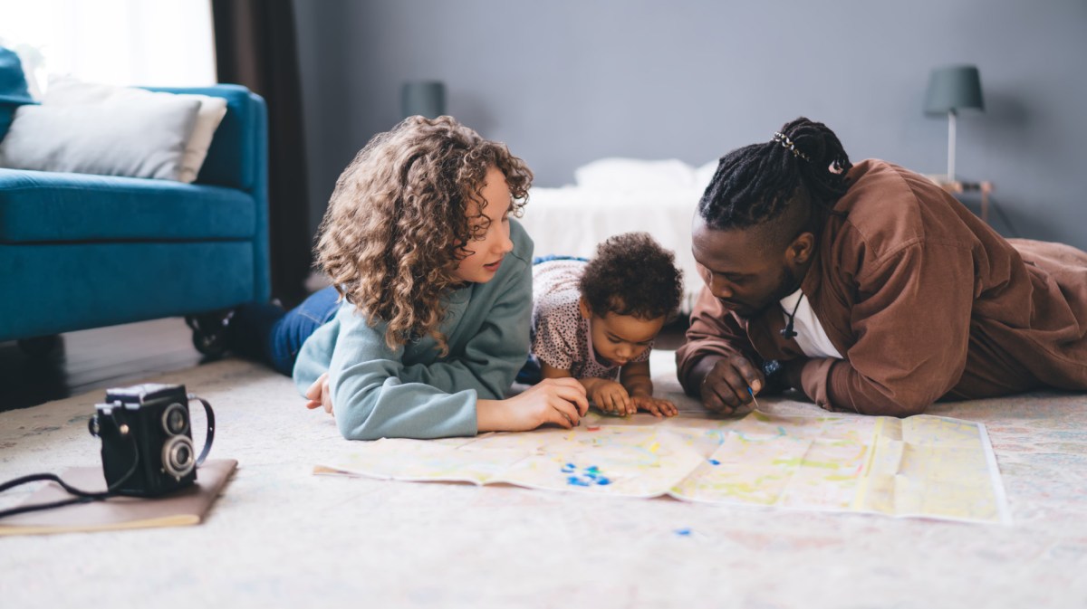 Mutter, Vater und Kleinkind liegen auf dem Fußboden und schauen eine Landkarte an.