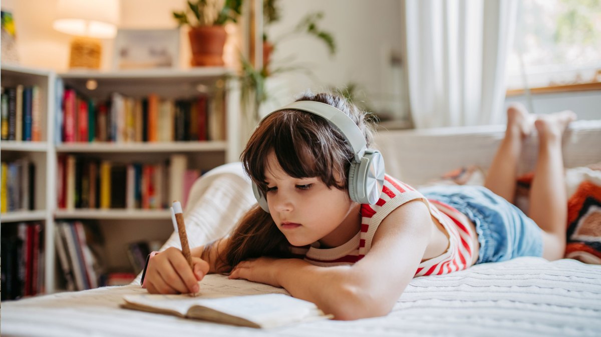 Mädchen liegt an einem warmen Tag drinnen auf ihrem bett, hört etwas über große Kopfhörer und schreibt in ein kleines Buch.