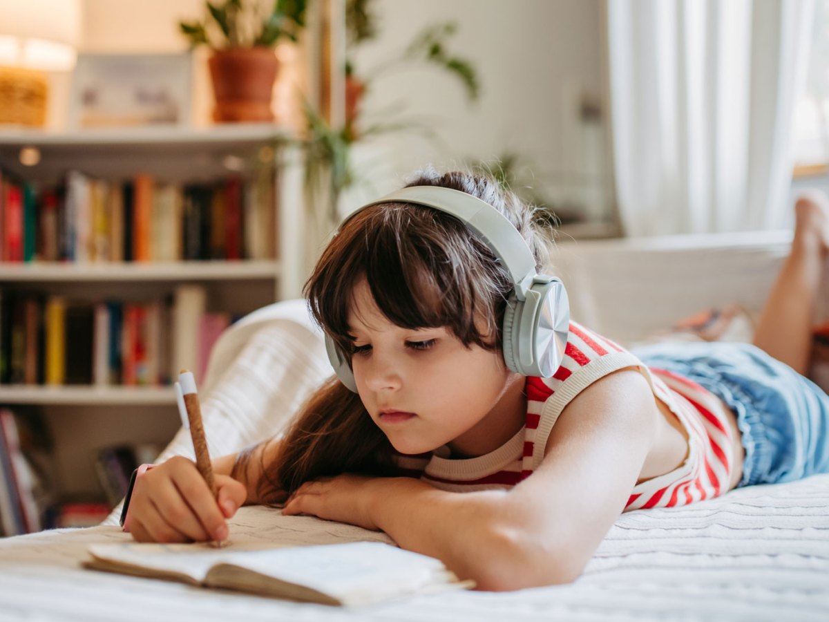 Mädchen liegt an einem warmen Tag drinnen auf ihrem bett, hört etwas über große Kopfhörer und schreibt in ein kleines Buch.