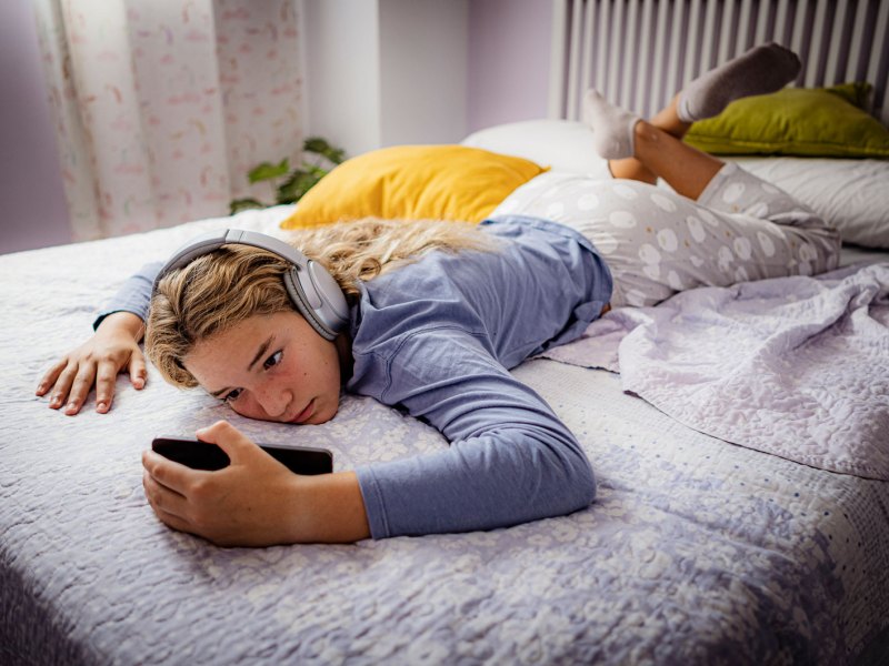 Mädchen im Teenageralter, liegt mit ihrem Handy und Kopfhörern faul auf ihrem Bett.