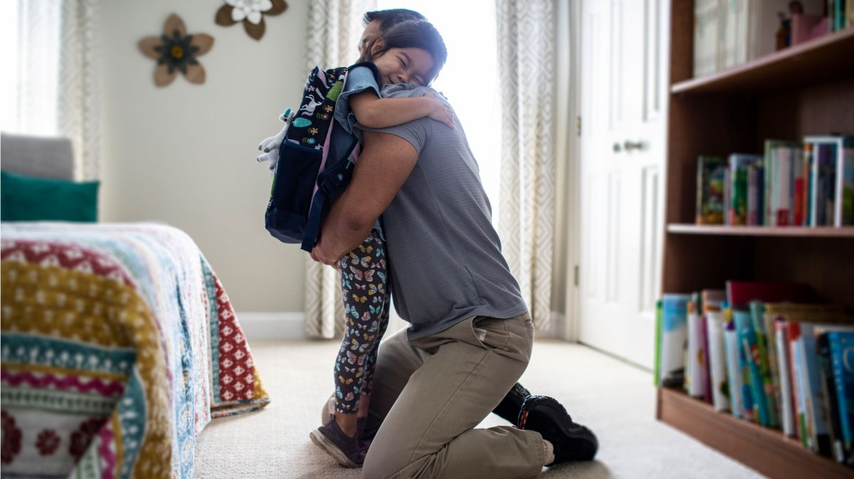 Vater umarmt seine Tochter, die einen Rucksack trägt, im Schlafzimmer.
