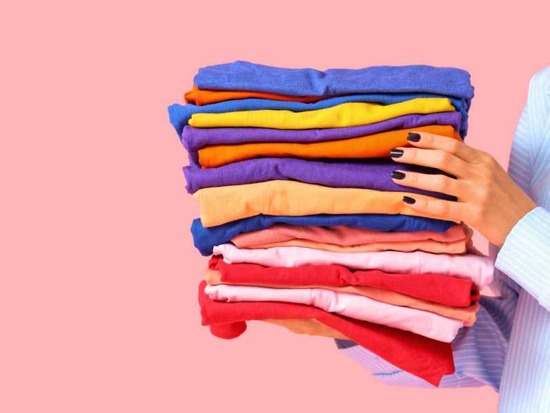 Junge Frau mit einem Stapel bunter Kleidung auf rosa Hintergrund, Nahaufnahme.