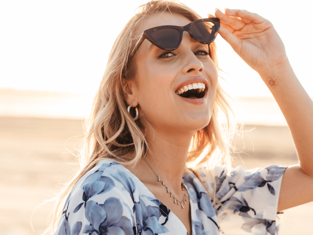 Frau mit Sonnenbrille an Strand l&auml;chelt in Kamera.