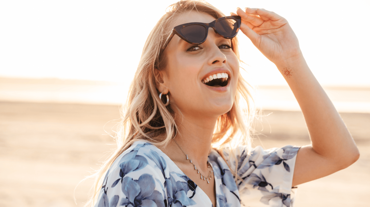 Frau mit Sonnenbrille an Strand lächelt in Kamera.