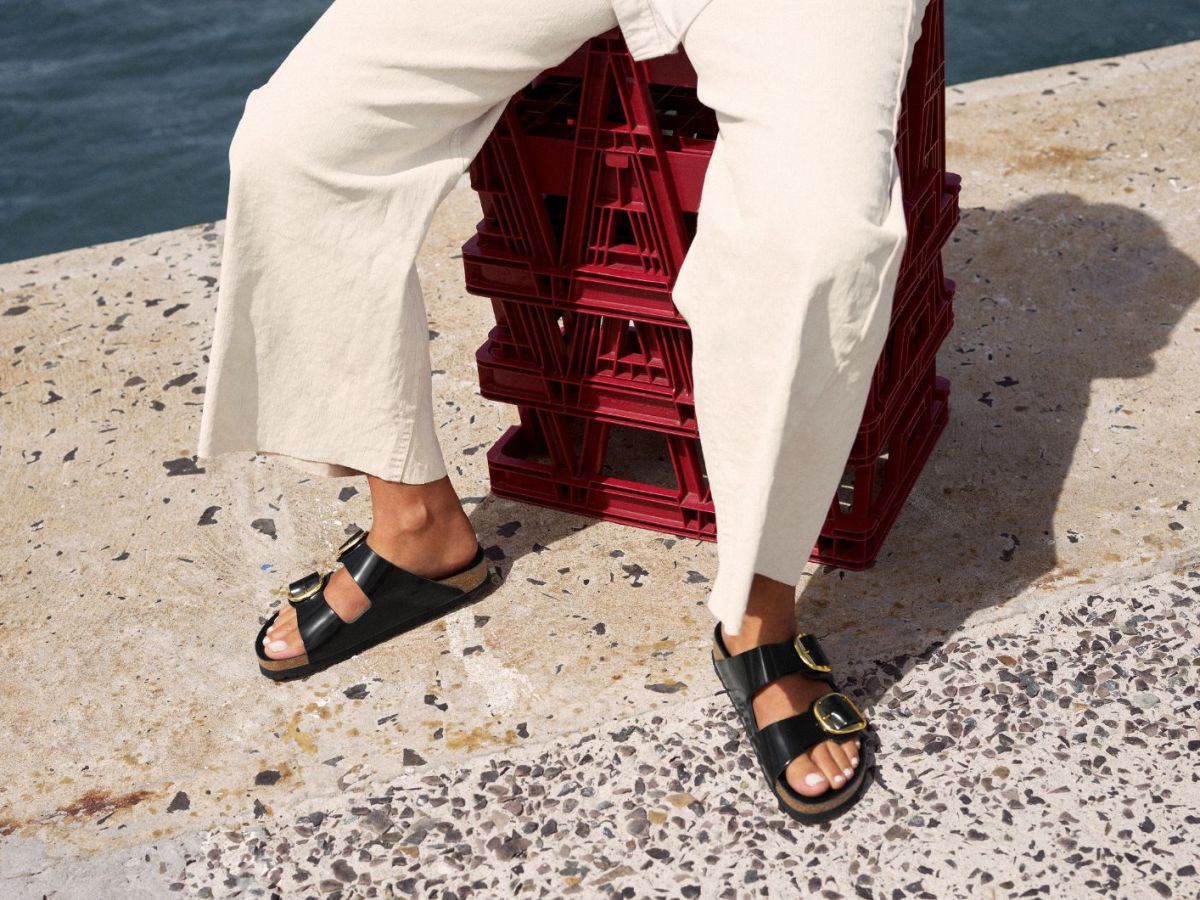 Frau in wei&szlig;er Jeans und schwarzen Birkenstock-Sandalen sitzt auf roten Kisten am Meer