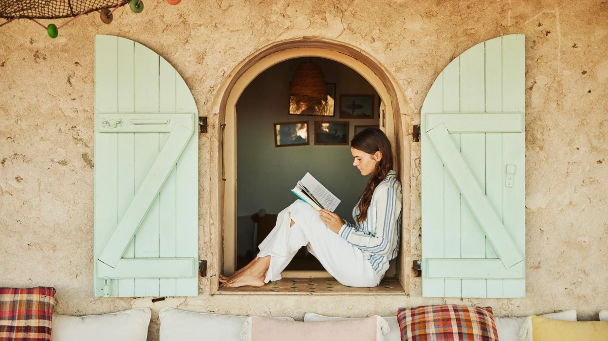 Junge Frau sitzt am Fenster und liest ein Buch, Urlaubsatmosphäre