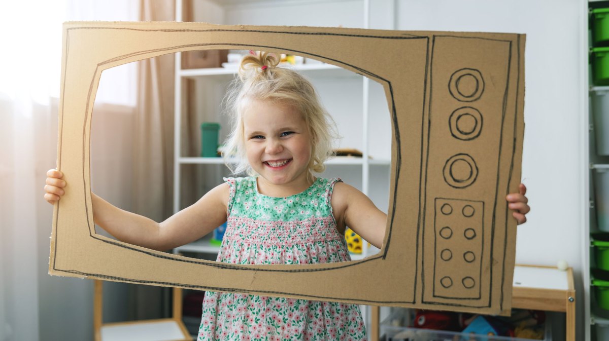 Mädchen hält einen aus Karton gebastelten Fernseher und schaut durch den Bildschirm.