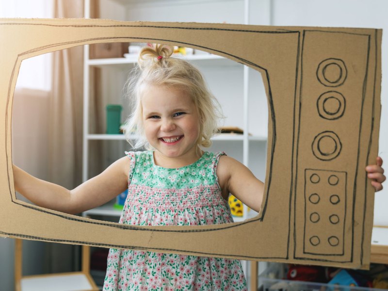 Mädchen hält einen aus Karton gebastelten Fernseher und schaut durch den Bildschirm.
