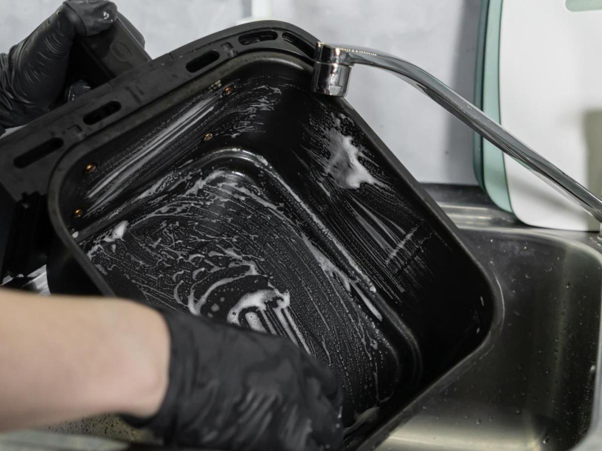 Person macht die Schublade eines Airfryers im Sp&uuml;lbecken mit einem Schwamm sauber.