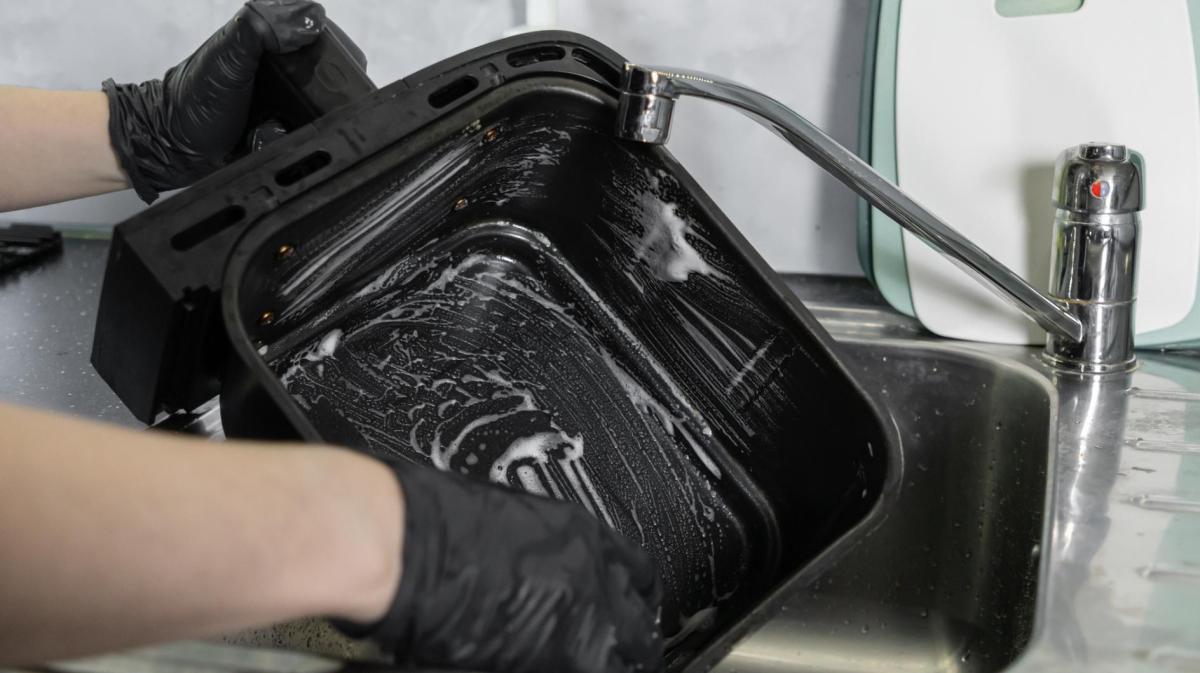 Person macht die Schublade eines Airfryers im Spülbecken mit einem Schwamm sauber.