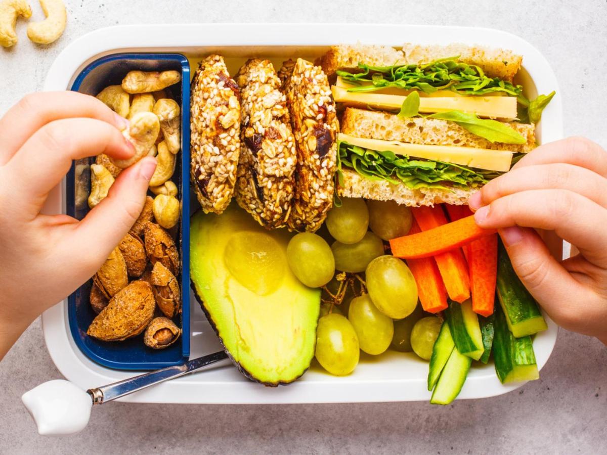 Kind sitzt vor einer Brotdose gef&uuml;llt mit Brot, Gem&uuml;se und Obst und isst davon.