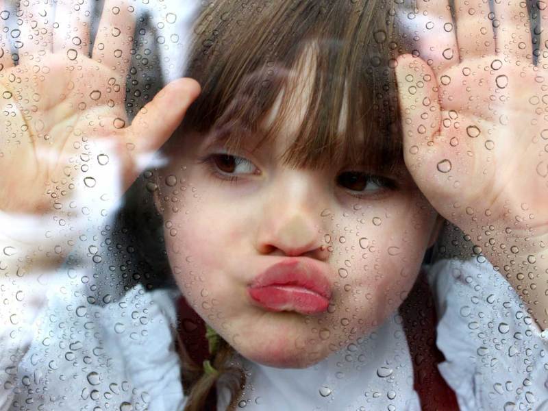 Mädchen drückt ihr Gesicht an die Fensterscheibe und schaut nach draußen, wo es augenscheinlich regnet.