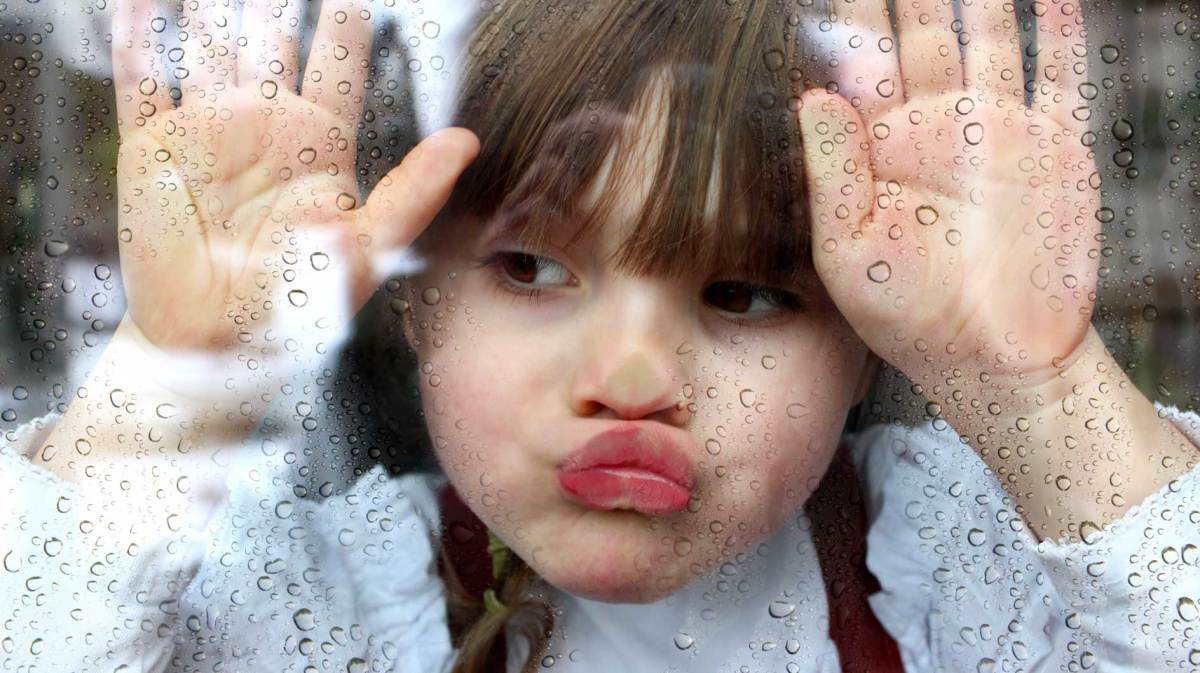 Mädchen drückt ihr Gesicht an die Fensterscheibe und schaut nach draußen, wo es augenscheinlich regnet.