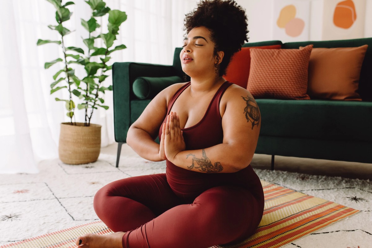 Dunkelhutige Frau mit weiblichen Kurven sitzt in einer entspannten Yoga-Position auf dem Wohnzimmerboden