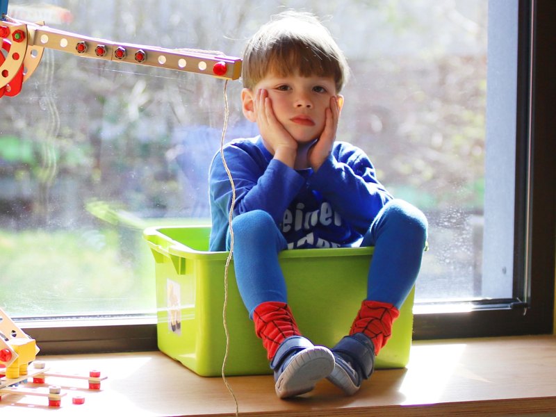 Kind sitzt in einer Spielkiste auf dem Fensterbrett und schaut gelangweilt.