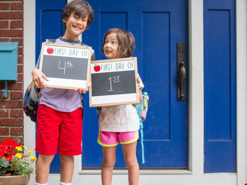 Großer Bruder und kleine Schwester vor der Haustür. Beide halten ein Schild in der Hand, auf dem zu lesen ist, welche Klassenstufen sie jetzt besuchen. Er startet die 4. Klasse, die kleine Schwester die 1.