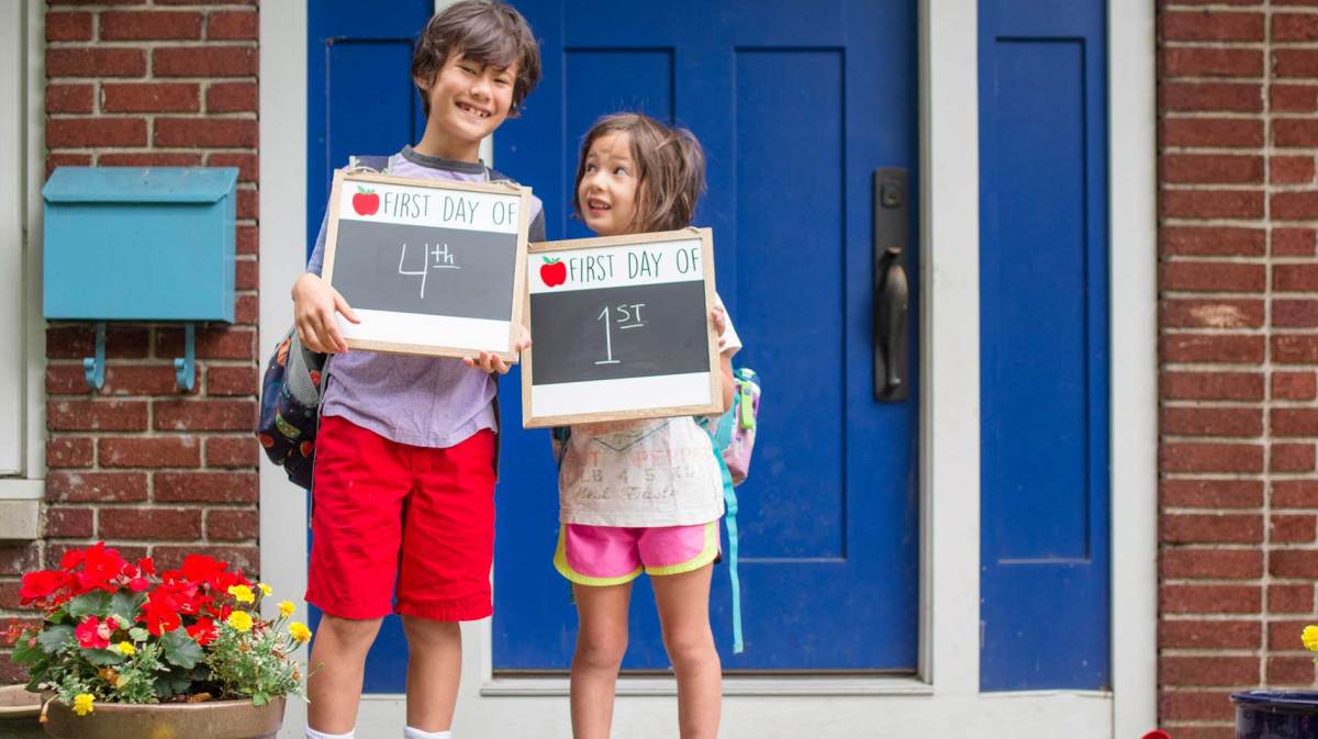 Großer Bruder und kleine Schwester vor der Haustür. Beide halten ein Schild in der Hand, auf dem zu lesen ist, welche Klassenstufen sie jetzt besuchen. Er startet die 4. Klasse, die kleine Schwester die 1.