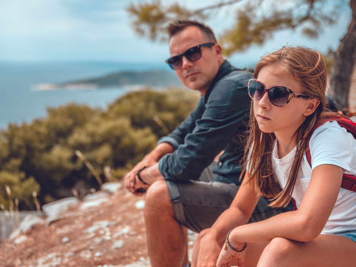 Vater und Tochter sitzen auf dem Gipfel eines Berges. Die Tochter sieht sehr genervt aus.