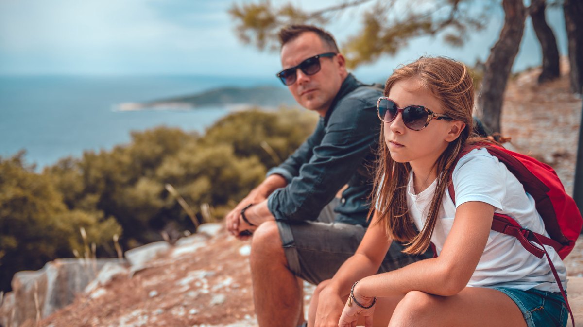 Vater und Tochter sitzen auf dem Gipfel eines Berges. Die Tochter sieht sehr genervt aus.