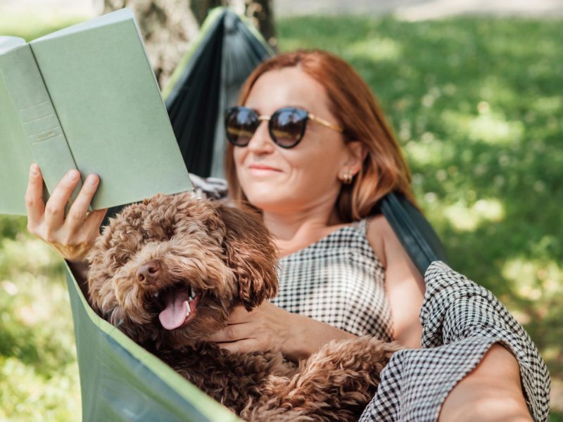 Frau liest in der Hängematte ein Buch und kuschelt mit ihrem Hund