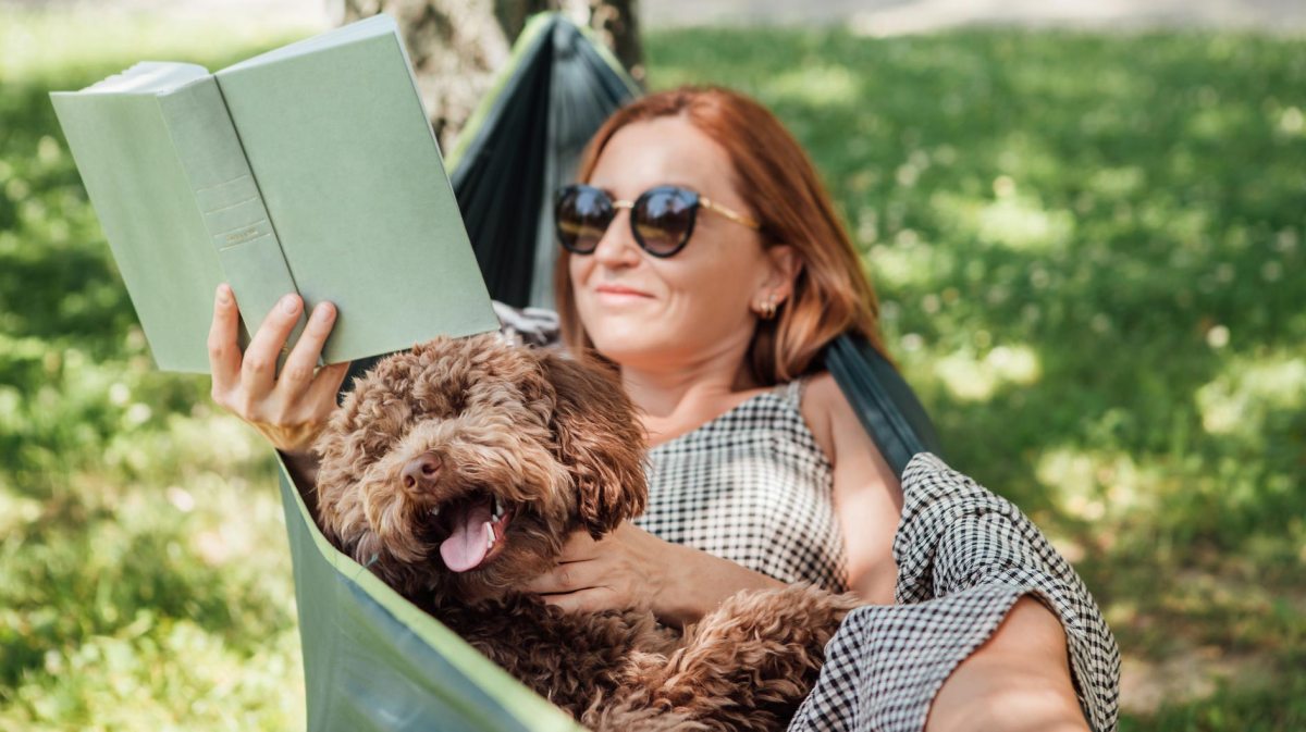 Frau liest in der Hängematte ein Buch und kuschelt mit ihrem Hund