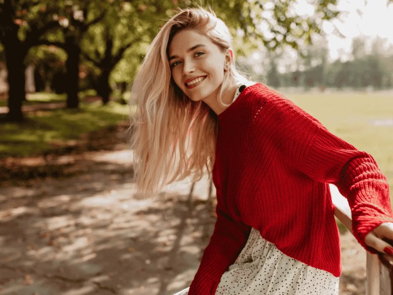 Blonde Frau in rotem Pulli lächelt in Kamera, hinter ihr ein herbstlicher Park.