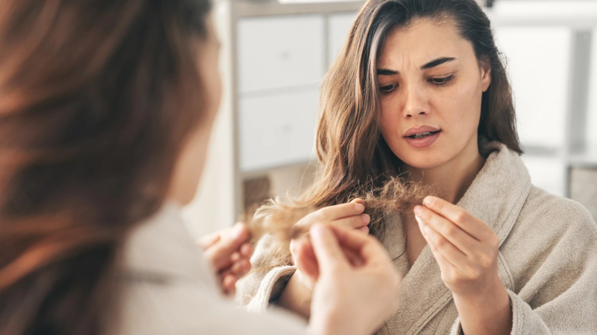 Junge Frau betrachtet ihre trockenen Haarspitzen im Spiegel.