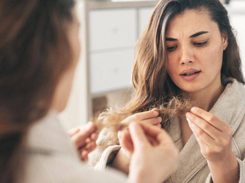 Junge Frau betrachtet ihre trockenen Haarspitzen im Spiegel.