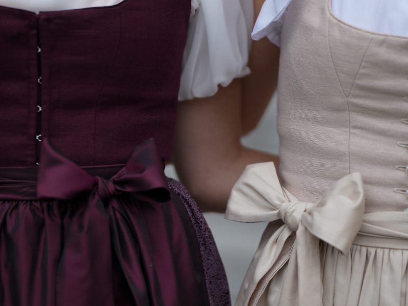 Zwei Frauen im Dirndl stehen eng nebeneinander, die Schleifen ihrer Schürzen sorgfältig vorne gebunden.