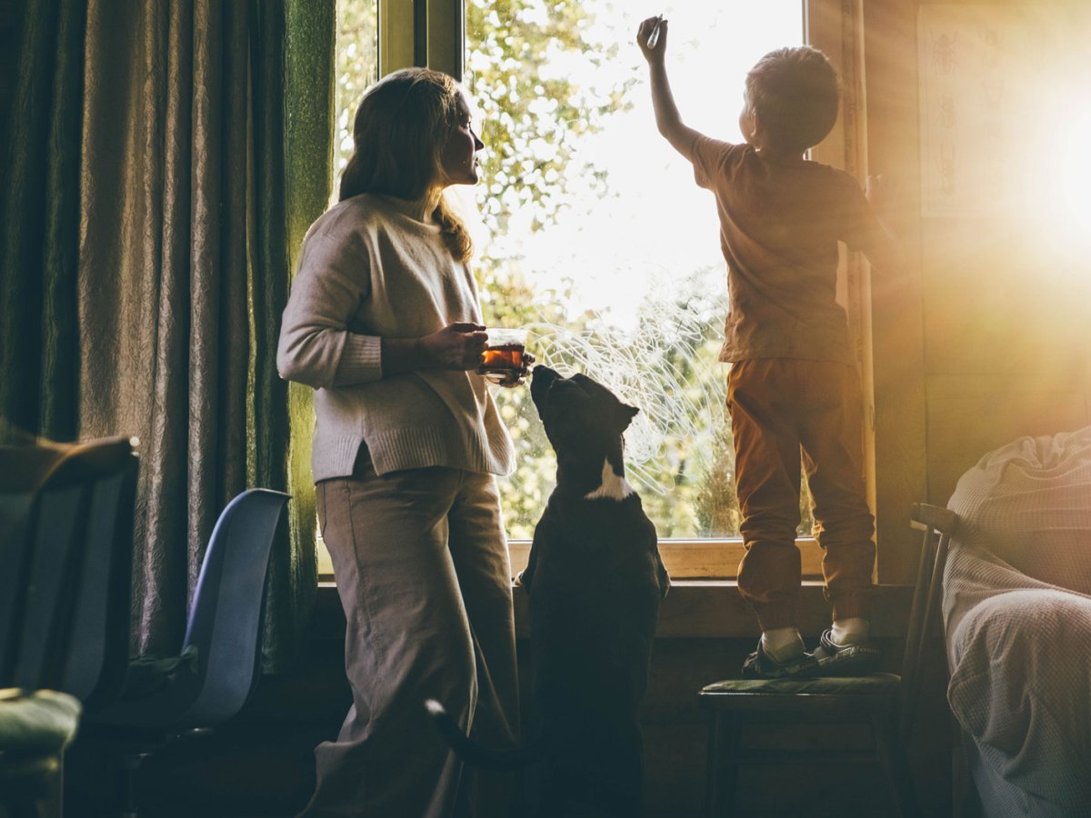 Mutter, Kind und Hund stehen in Pyjamas morgens am Fenster, die Sonne geht gerade auf und schauen nach draußen.