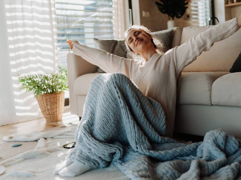 Frau sitzt in einer gemütlichen Wohnung, mit einer Decke auf ihrem Schoß