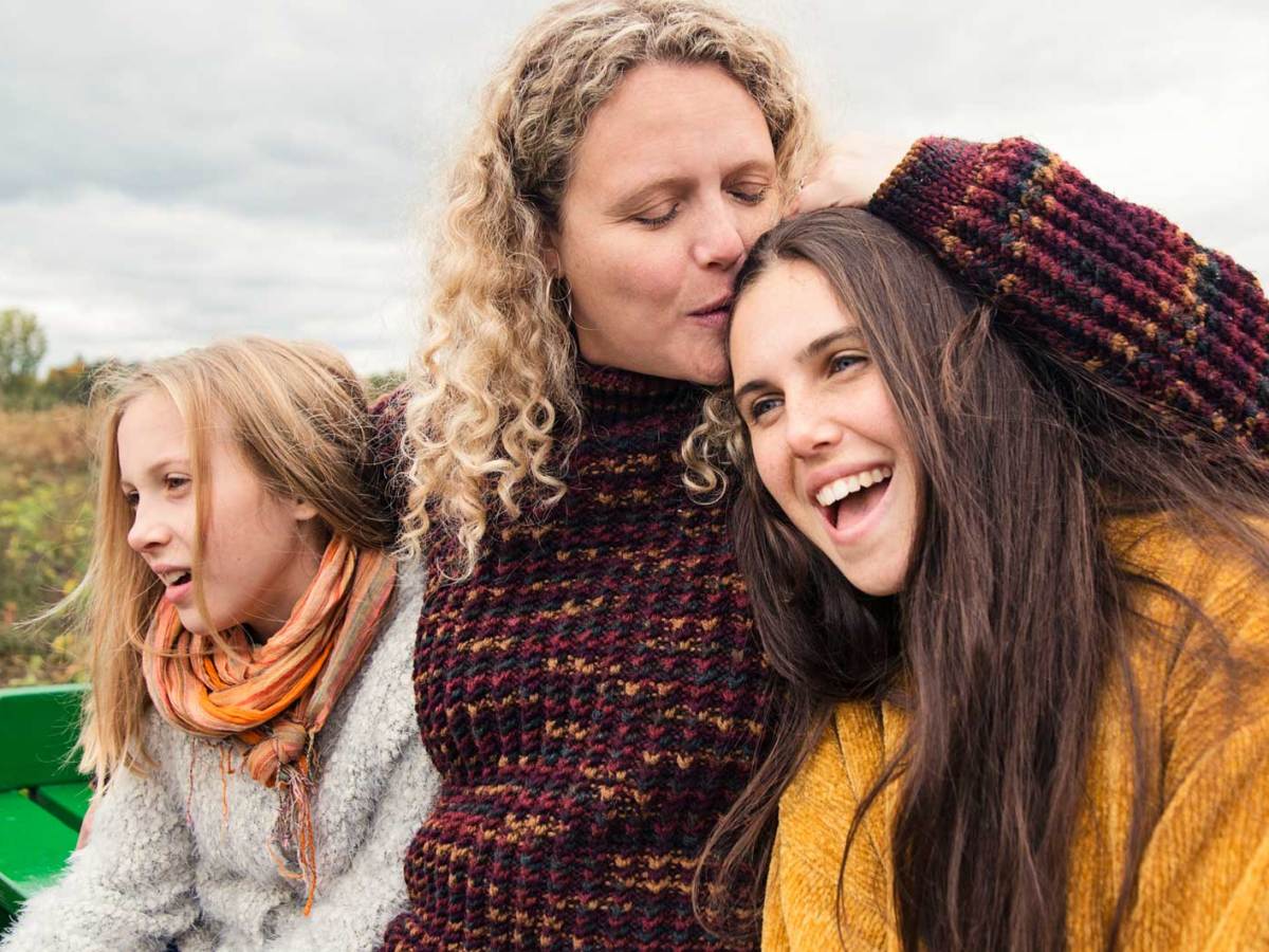 Mutter und zwei T&ouml;chter tragen warme Pullis und sitzen an einem bew&ouml;lkten Herbsttag auf einer Bank in der Natur.