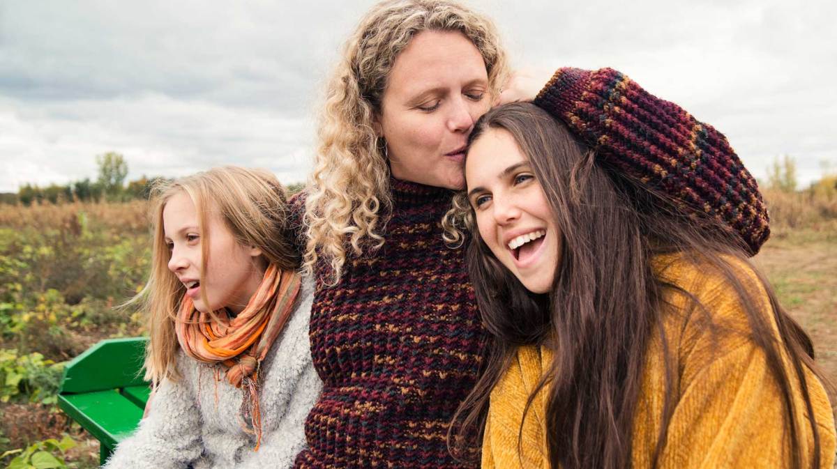 Mutter und zwei Töchter tragen warme Pullis und sitzen an einem bewölkten Herbsttag auf einer Bank in der Natur.