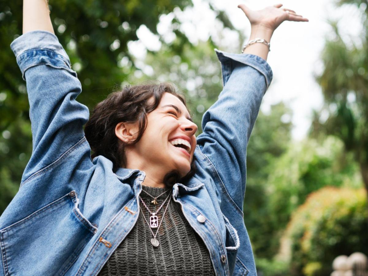 Junge Frau mit braunen Haaren und Jeansjacke wirft die Arme in die Luft und freut sich.
