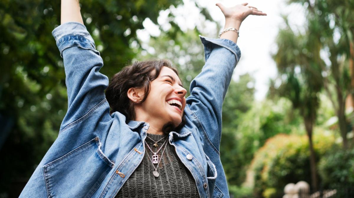 Junge Frau mit braunen Haaren und Jeansjacke wirft die Arme in die Luft und freut sich.