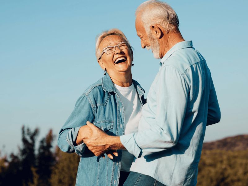 Älteres Paar steht in Feld, die Frau lacht beherzt, der Mann hält sie an den Händen.