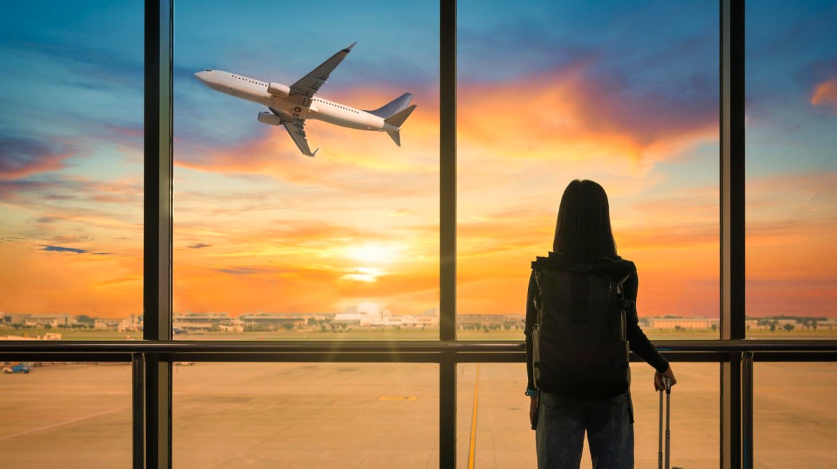 Frau steht mit Koffer am Flughafen und beobachtet aufsteigendes Flugzeug bei Sonnenuntergang