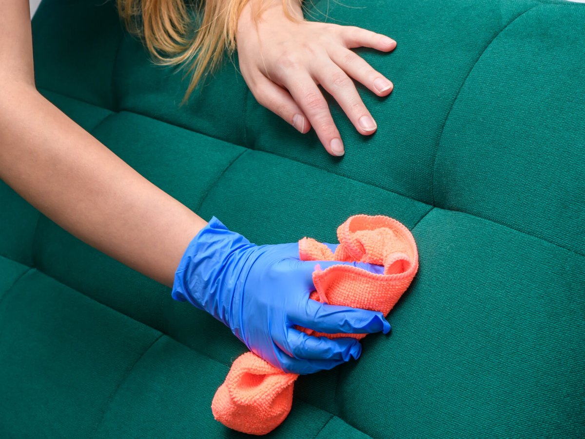 Junge Frau reinigt mit orangefarbenem Tuch und blauen Einmalhandschuhen gründlich ein grünes Polstersofa.