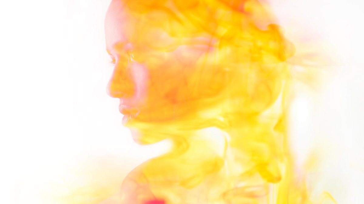 Profil einer jungen Frau, deren Gesicht von leuchtenden Gelb- und Orangenschleiern weich umhüllt wird.