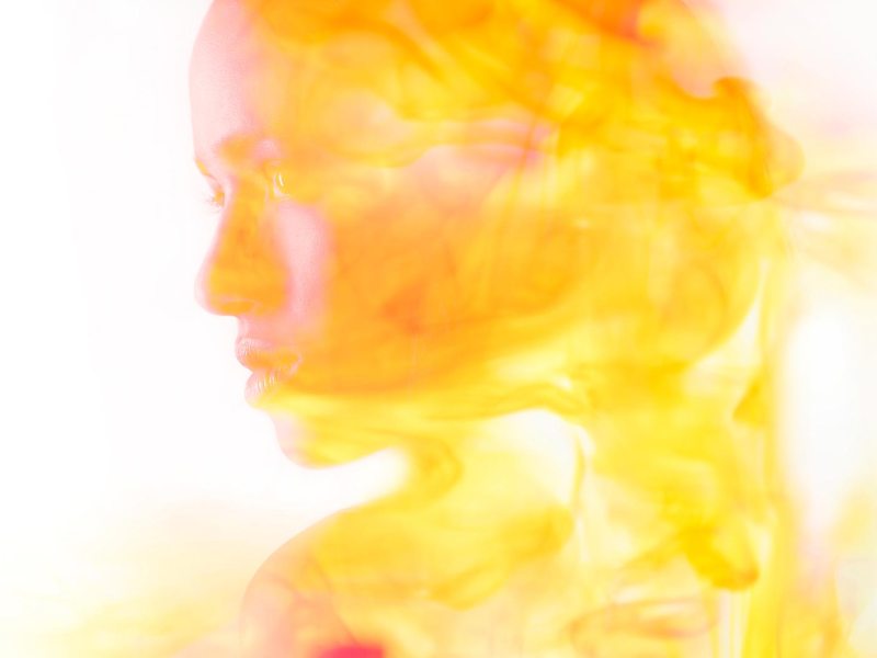 Profil einer jungen Frau, deren Gesicht von leuchtenden Gelb- und Orangenschleiern weich umhüllt wird.