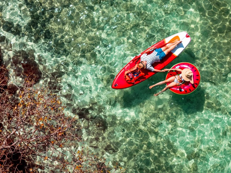 Ein junges Paar entspannt im klaren türkisgrünen Wasser, sie auf einer Wassermelonen-Luftmatratze, er auf einem roten SUP