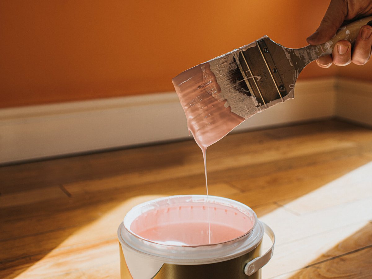 Eine Hand h&auml;lt einen tropfenden Pinsel mit rosafarbener Farbe &uuml;ber einem offenen Farbeimer auf hellem Holzboden.