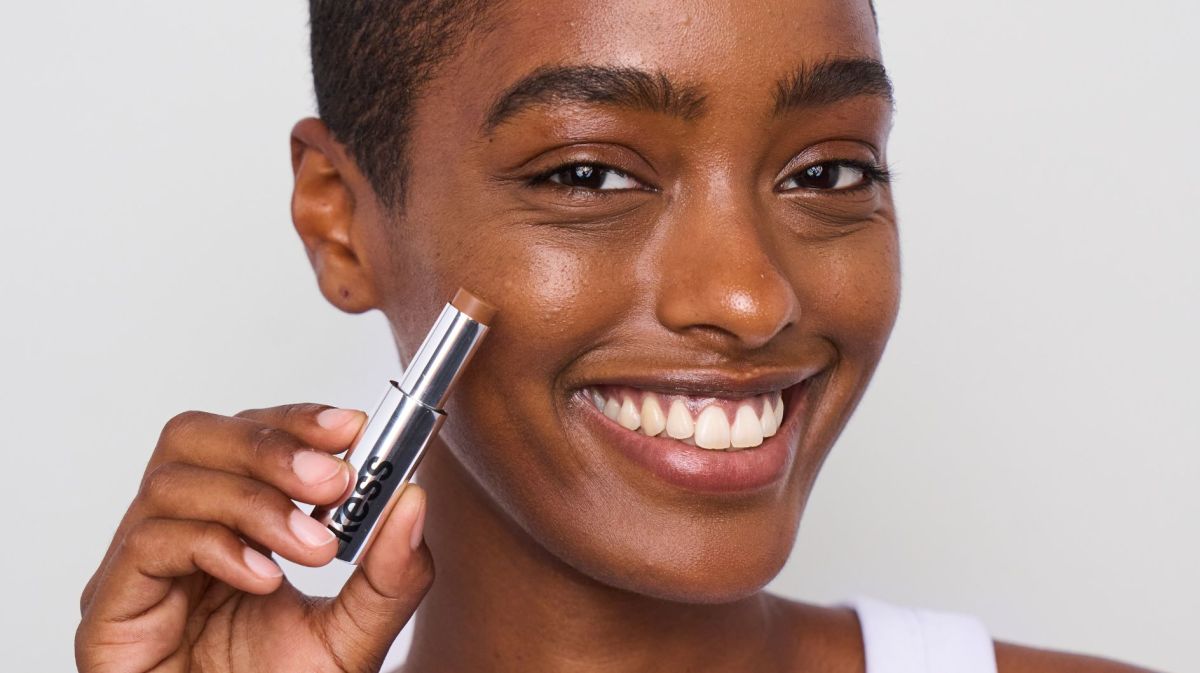 Junge, lächelnde Frau mit kurzen Haaren hält Concealer an die Wange