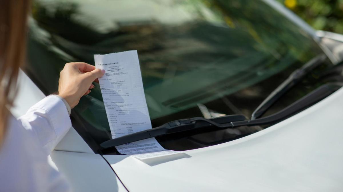 Frau zieht ein Knöllchen unter den Scheibenwischern hervor.