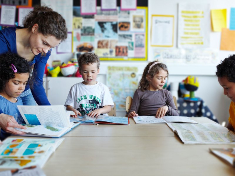 Gruppe Erstklässler sitzt mit Büchern an einem großen, runden Tisch, die Lehrerin beugt sich über eines der Kinder, um zu helfen.