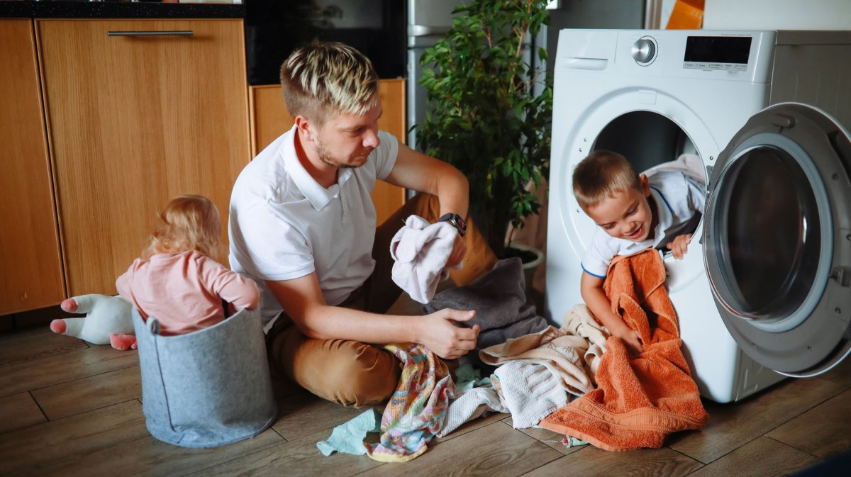 Vater sitzt vor der Waschmaschine auf dem Boden. Sein Sohn schaut aus der Wäschetrommel und die Tochter sitzt daneben in einem Stoffsack.