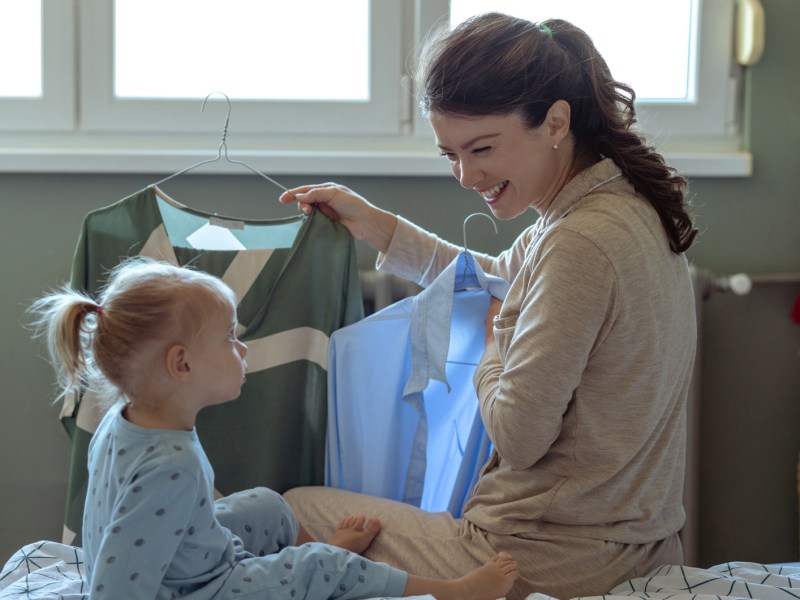 Frau, die mit ihrem Kind auf dem Bett sitzt und überlegt, was sie anzieht.